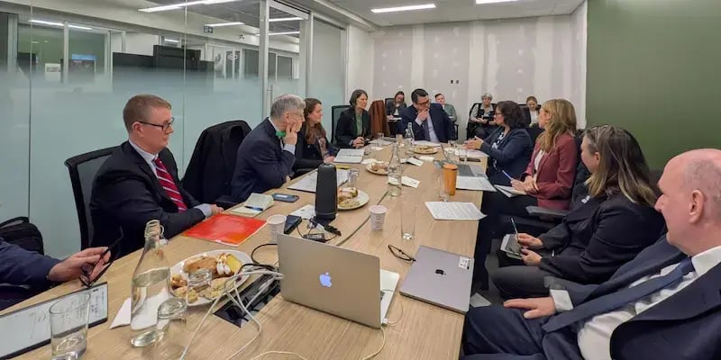 Professional group in business attire sits around a long wooden boardroom table. They are engaged in a meeting with laptops, notebooks, and water bottles present. The setting is a modern office with glass walls and a green accent wall. Some participants take notes while others listen intently.