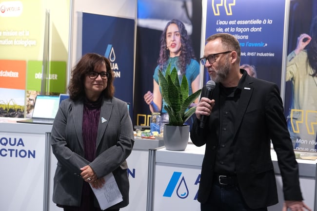 Frédéric Vincent parle dans un micro au stand d'AquaAction. Soula Chronopoulos se tient à ses côtés. Derrière eux, des bannières AquaAction traitent de la transformation écologique et de l'eau, avec une plante Sansevieria posée sur le comptoir.