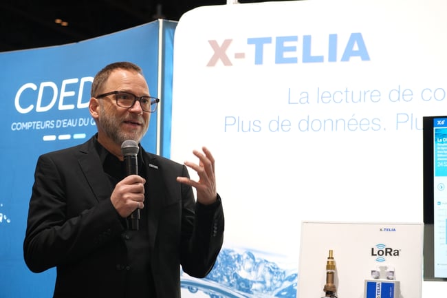 Frédéric Vincent en veston noir parle au micro et gesticule lors d'une présentation. Derrière lui, on voit les enseignes X-TELIA et CDED, ainsi qu'un présentoir de technologie LoRa avec du matériel et un écran de données.