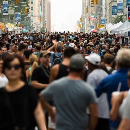 A busy urban street filled with a large crowd of people attending an outdoor event, with market stalls and city buildings in the background, symbolizing urban life and social gatherings.