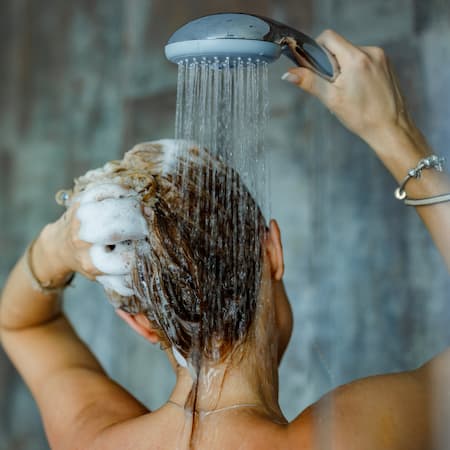 A woman washing her hair under a running shower, with shampoo lather in her hair, representing daily water consumption and personal hygiene.