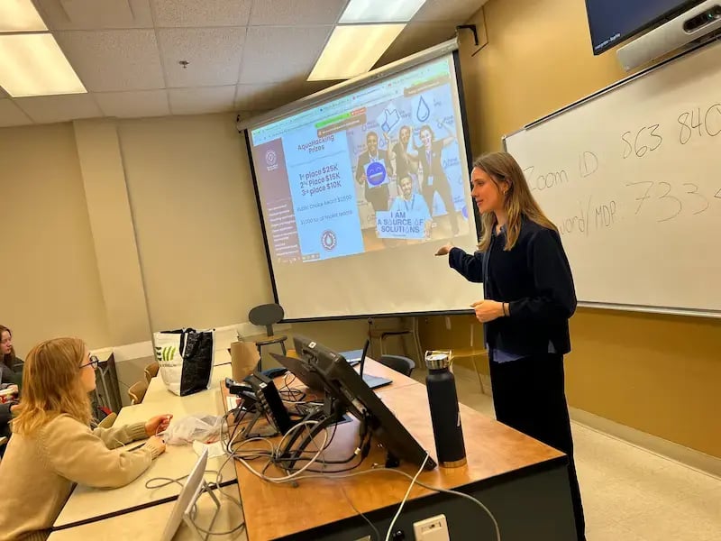 A young woman with long brown hair, identified as Kenza, stands in a classroom presenting the AquaHacking Great Lakes 2026 challenge. She is wearing a dark blue corduroy shirt and gestures toward a large projection screen displaying "AquaHacking Prizes." The screen lists 1st place as $25K, 2nd place as $15K, and 3rd place as $10K, alongside a photo of smiling participants holding a sign that says "I AM A SOURCE OF SOLUTIONS." In the foreground, a student with blonde hair sits at a desk listening intently.