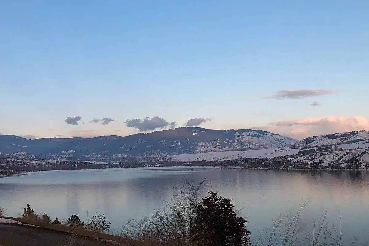 Une vue sereine du lac Okanagan avec des montagnes enneigées en arrière-plan, reflétant le ciel bleu calme et les nuages épars au crépuscule.