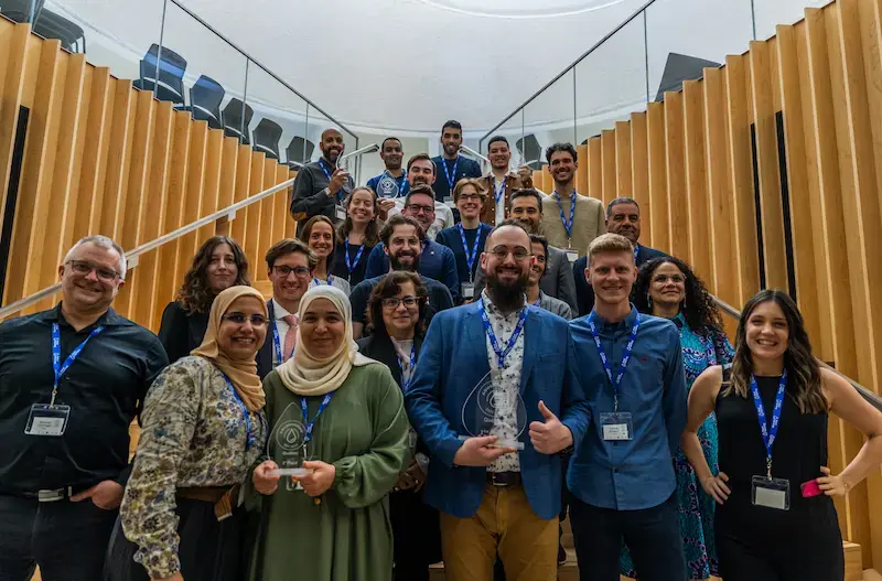 A large group of smiling winners and participants pose on a modern wooden staircase during AquaHacking Québec 2023. Several individuals in the front and back rows proudly hold clear glass trophies. The group is diverse, wearing professional attire and blue event lanyards.
