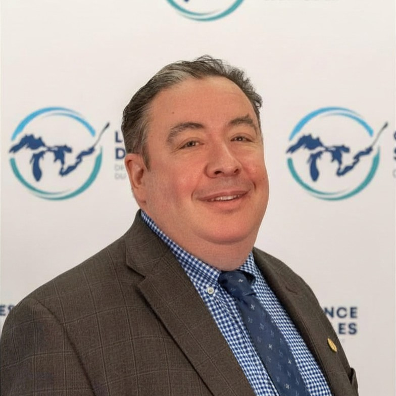 John Altenberg smiles in a portrait wearing a dark grey patterned suit jacket, blue-and-white patterned button-down shirt, and patterned blue tie. He stands before a white backdrop repeating a blue circular logo with Great Lakes outlines.