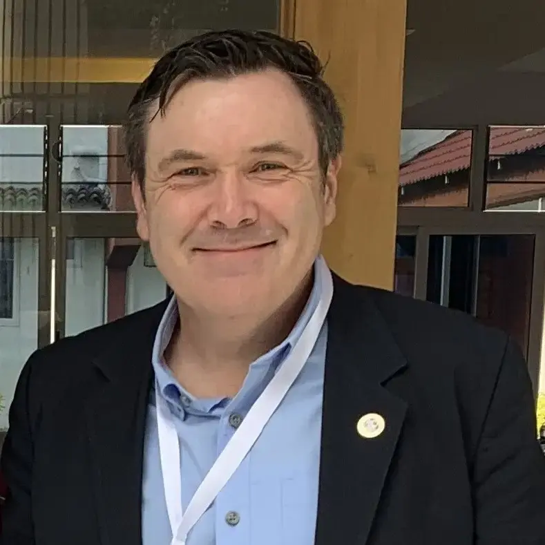 Mike McKay smiles in a headshot portrait, wearing a light blue dress shirt and a dark blazer with a round gold lapel pin. A white conference lanyard hangs around his neck. He has short dark hair and a warm expression in front of a windowed backdro