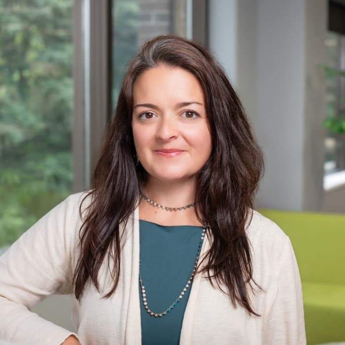 Delaney Keating offers a gentle smile in a professional portrait. She wears a teal top, a off-white cardigan, a beaded choker, and a longer beaded necklace. She has long, dark wavy hair. Trees are visible through a large window in the background.