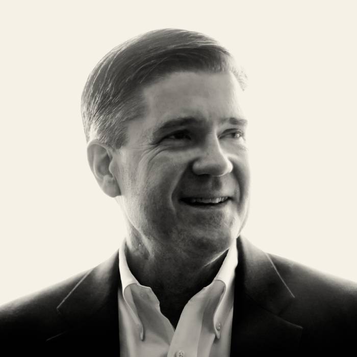 John Austin smiles while looking to the right in a black-and-white portrait. He wears an open-collar white dress shirt and a dark suit jacket. Strong backlighting highlights his short hair, suit silhouette, and profile against a bright white background.