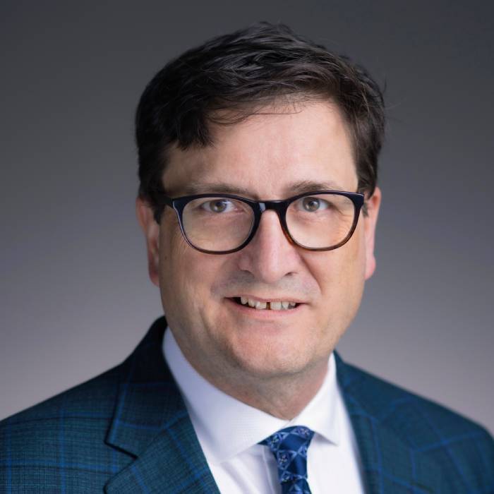 Mark Schrupp wears black-framed glasses, a blue plaid suit jacket, a white dress shirt, and a patterned blue tie. He has dark brown hair and offers a slight smile in a studio headshot against a neutral grey background.