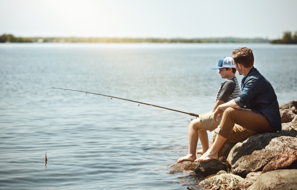 A man and a young boy sit barefoot on large rocks at the water's edge, fishing on a sunny day. They are side-by-side, holding a fishing rod out over the calm lake or reservoir.