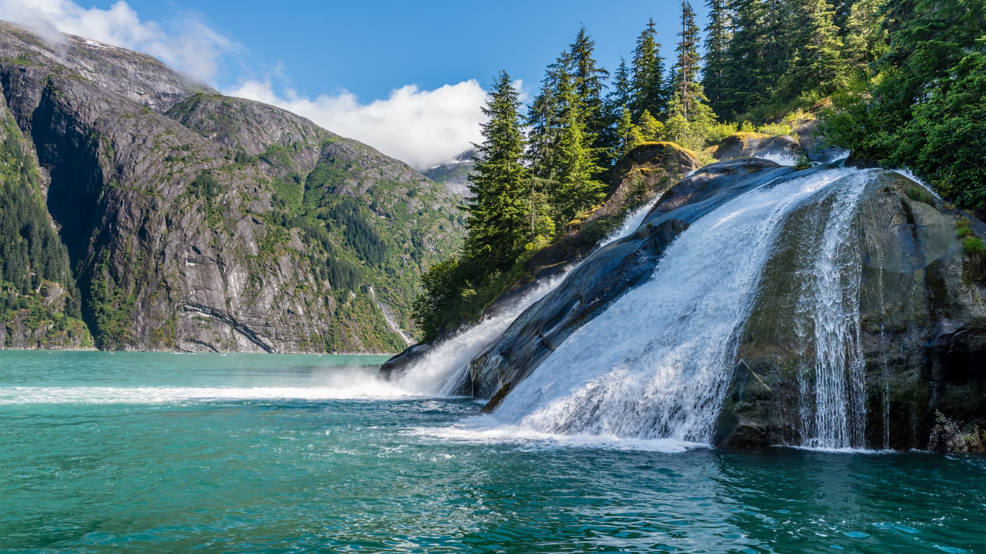 A powerful waterfall cascades into a turquoise lake surrounded by lush green forests and rugged mountains.