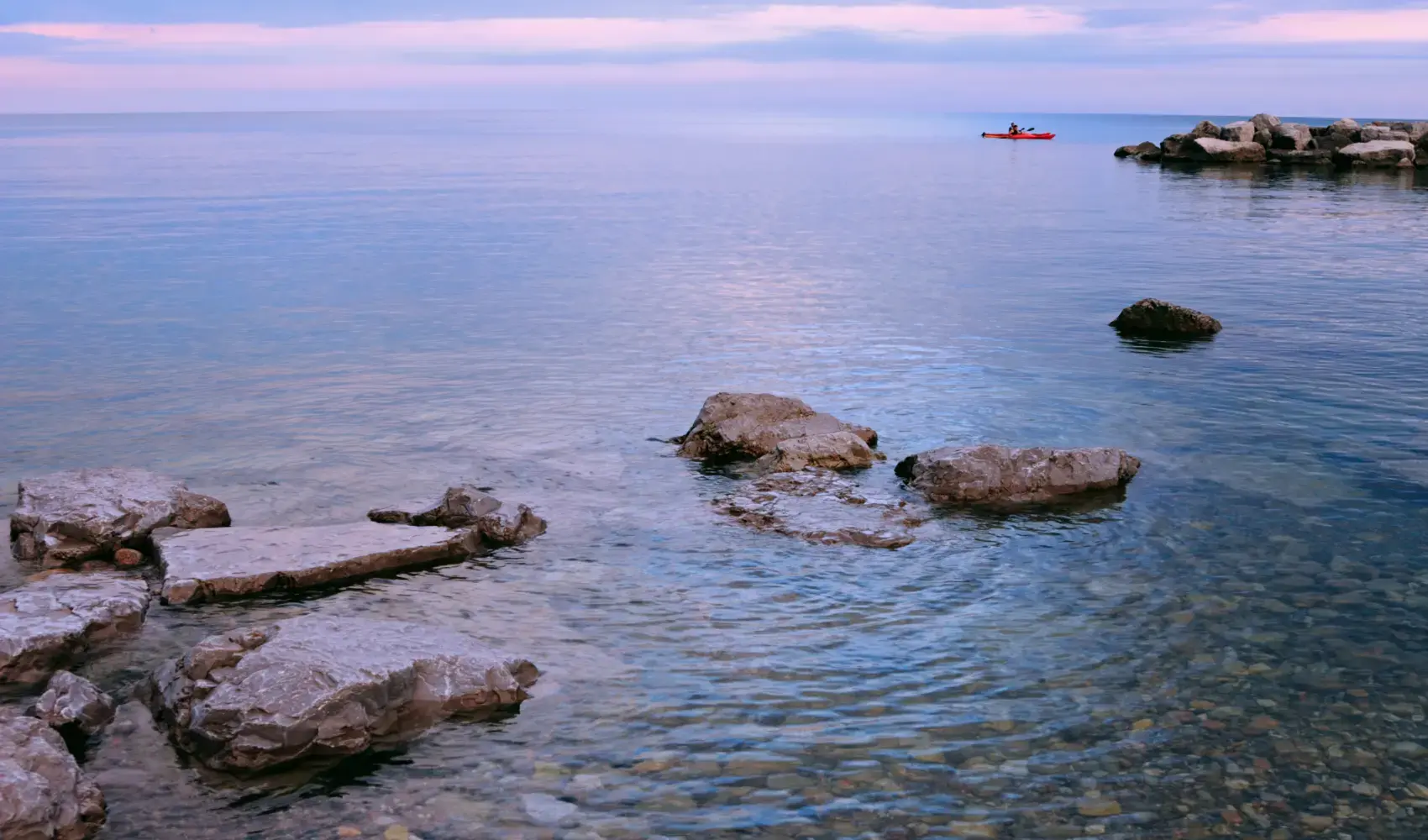 A tranquil scene at Lake Ontario with a kayak gently floating past rocky outcrops, under a soft evening sky.