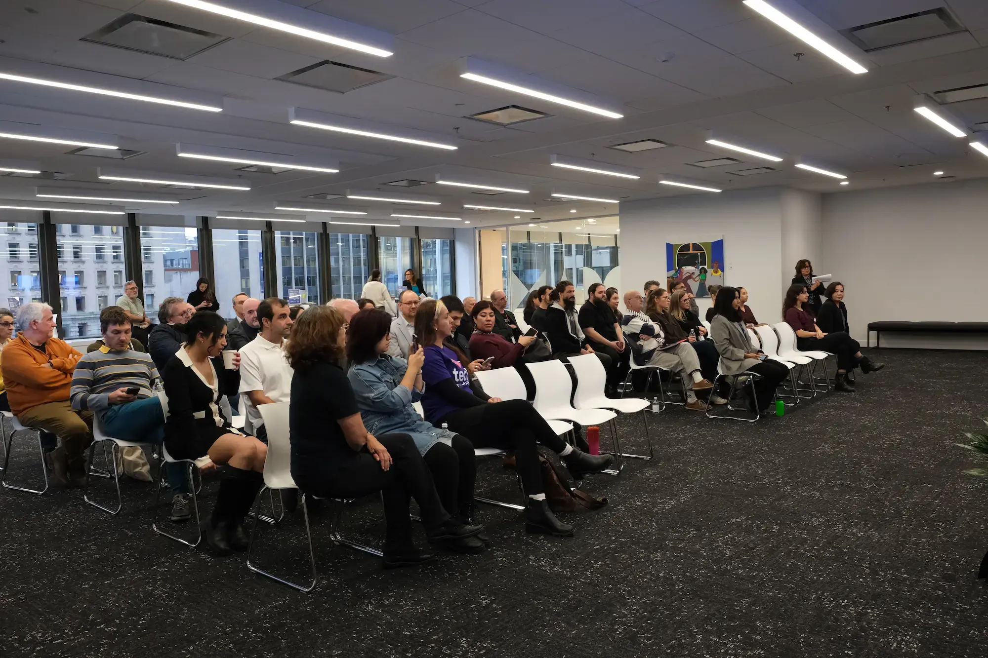 Une vue d'ensemble d'un public diversifié assis dans des rangées de chaises blanches dans un espace événementiel moderne à aire ouverte. De grandes fenêtres en arrière-plan montrent des bâtiments de la ville, et la salle est éclairée par de longs plafonniers rectangulaires. Une vue d'ensemble d'un public diversifié assis dans des rangées de chaises blanches dans un espace événementiel moderne à aire ouverte. De grandes fenêtres en arrière-plan montrent des bâtiments de la ville, et la salle est éclairée par de longs plafonniers rectangulaires.