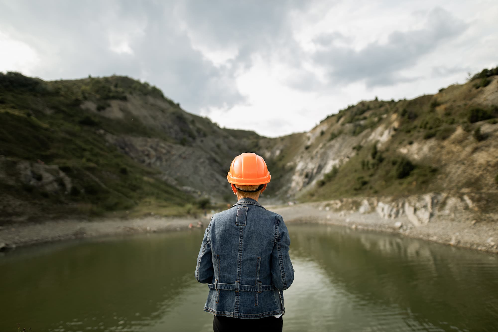Person wearing an orange hard hat and denim jacket inspecting a body of water in a quarry or natural site, with hills in the background.