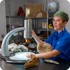 A woman with green hair uses a digital microscope to examine specimens in a science lab. On the desk are an ammonite fossil and a dinosaur skull. The background features shelves with a globe, a volcano model, and various geological samples.